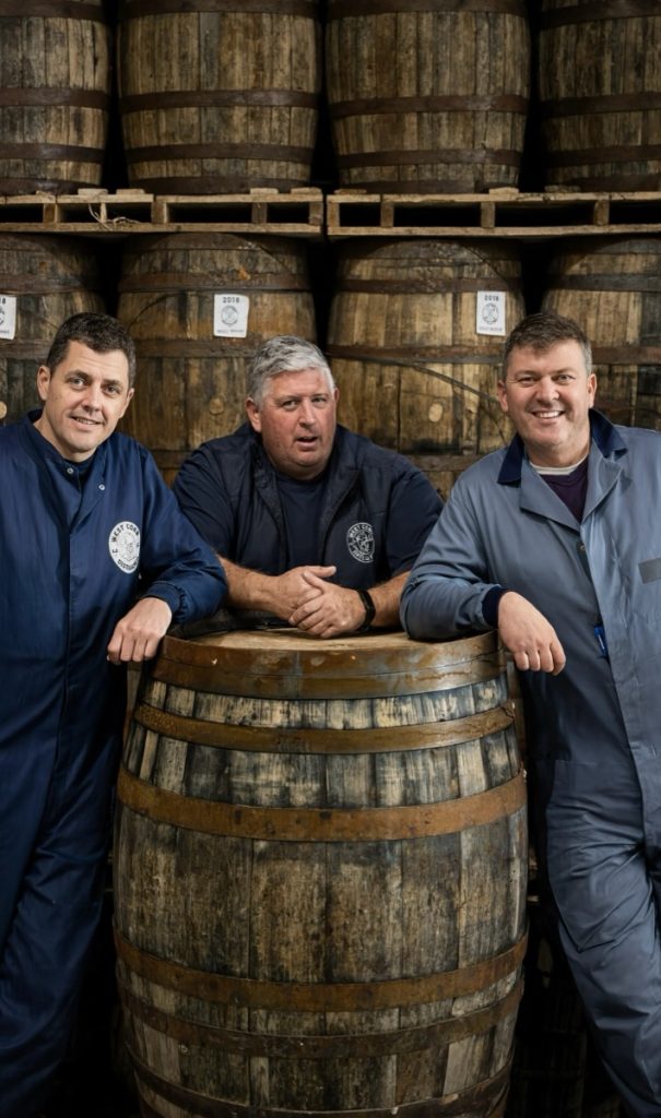 Three members of the West Cork Distillery team leaning on a whiskey barrel in the warehouse