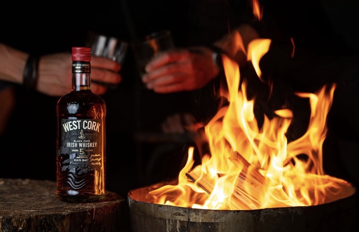 A bottle of West Cork Black Cask beside a fire pit, with drams poured for friends